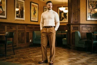 Homme élégant en tenue rétro 2026 avec pantalon large caramel et pull col montant ivoire dans un salon vintage aux tons ambrés